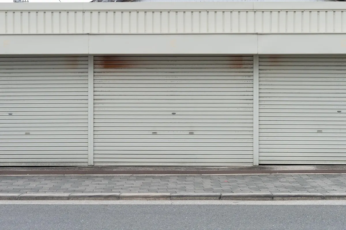 Trois rideaux métalliques gris clair baissés sous un bandeau de commerces, au bord d'un trottoir