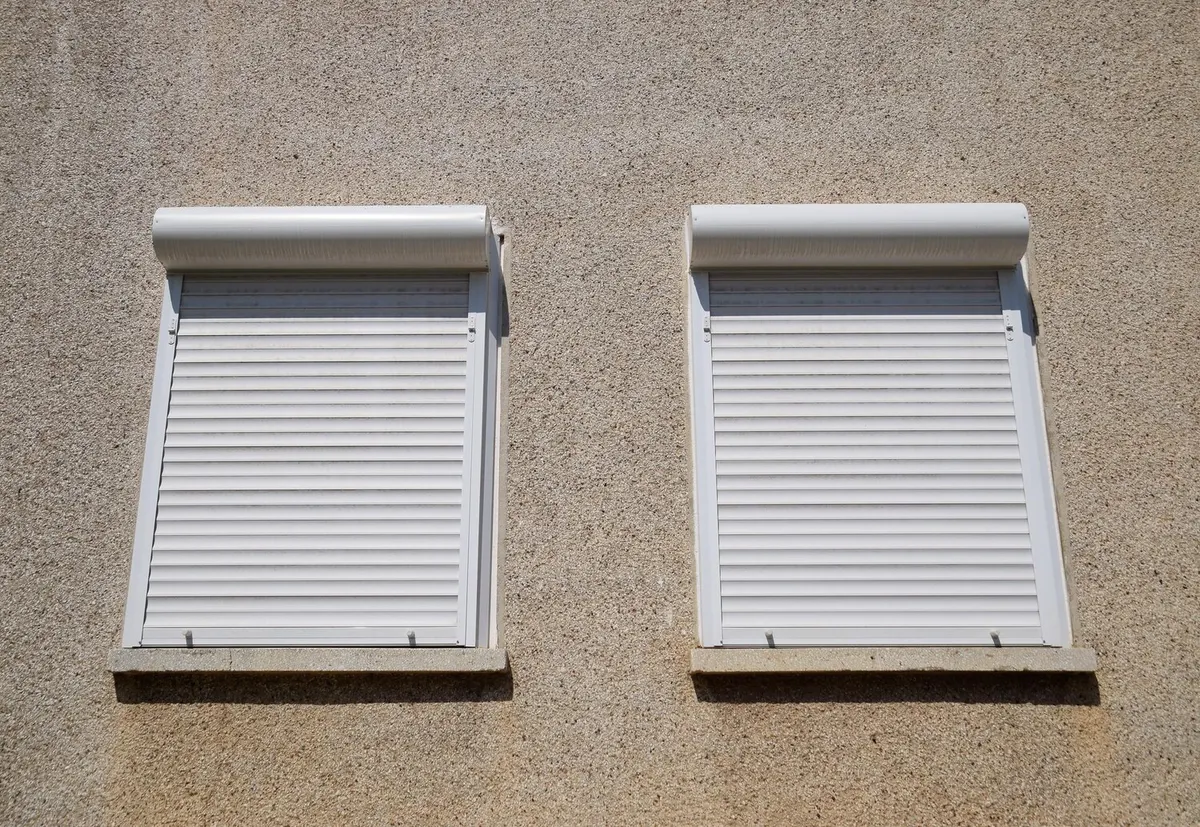 Deux volets roulants blancs à projection à l'italienne, tabliers inclinés vers l'extérieur, sur une façade crépie beige